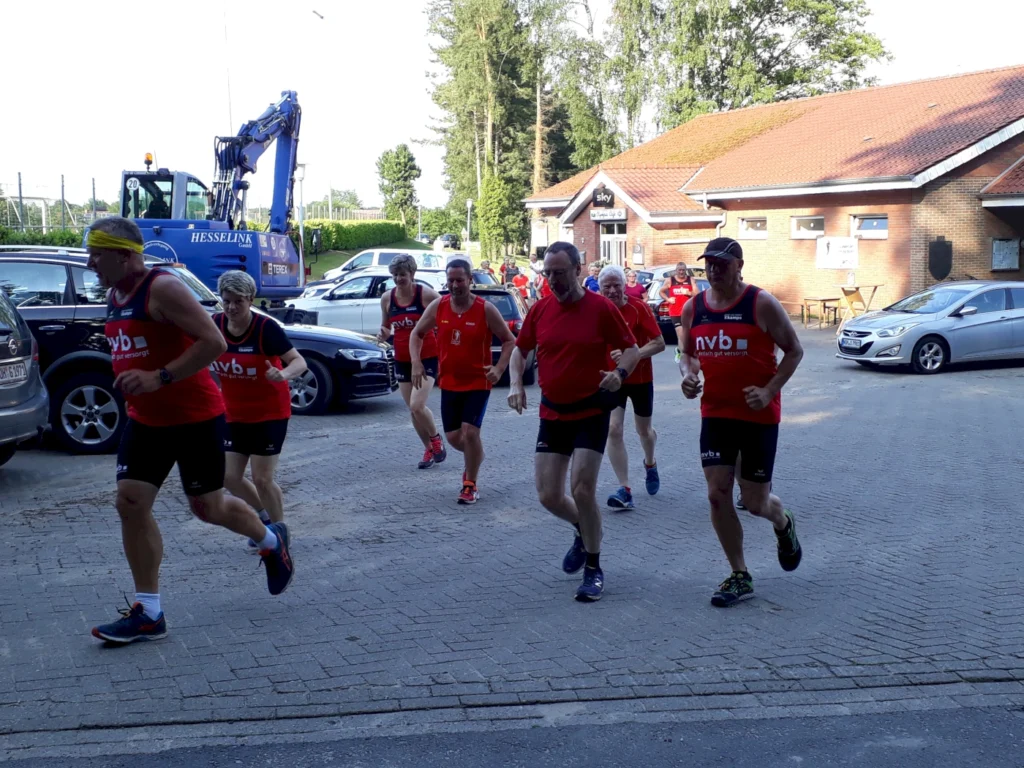 Gruppe von Läufern in roten Trikots, die auf einem gepflasterten Parkplatz joggen. Im Hintergrund sind Autos und ein Bagger sichtbar, während die Umgebung von Bäumen umgeben ist.