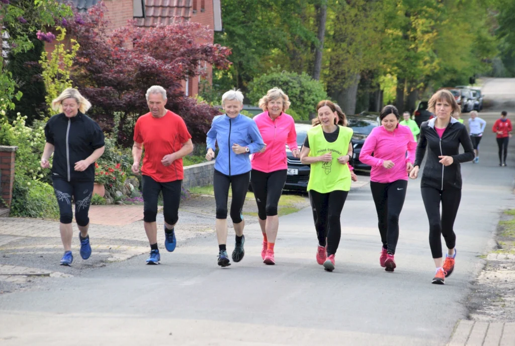 Gruppe von sieben joggenden Menschen auf einer Straße, umgeben von grüner Natur und Wohnhäusern. Die Teilnehmer tragen bunte Sportkleidung und scheinen gemeinsam an einem Fitness-Event oder Lauftraining teilzunehmen.