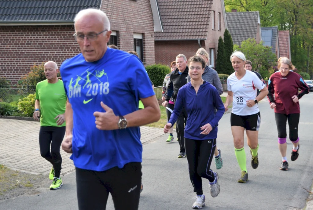 Gruppe von Läufern, darunter Männer und Frauen unterschiedlichen Alters, die auf einer Straße joggen. Im Vordergrund ein älterer Mann in einem blauen Shirt mit der Aufschrift "BM 2018". Die Umgebung zeigt Wohnhäuser und eine grüne Landschaft.