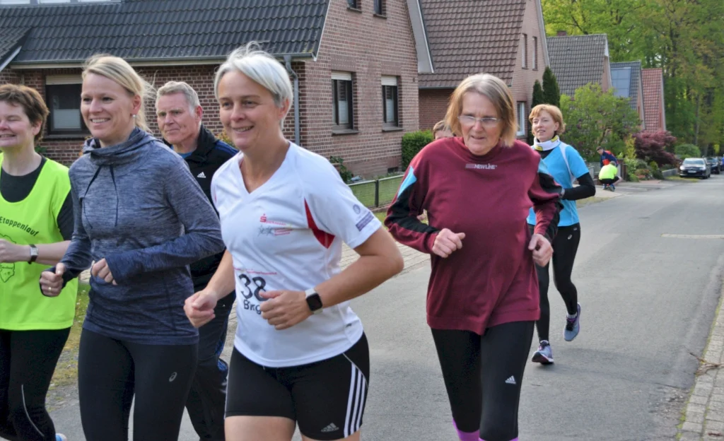 Gruppe von Läufern beim Etappenlauf auf einer Straße, verschiedene Altersgruppen und Fitnessniveaus, fröhliche Gesichter und sportliche Kleidung, umgeben von Wohnhäusern und Bäumen.
