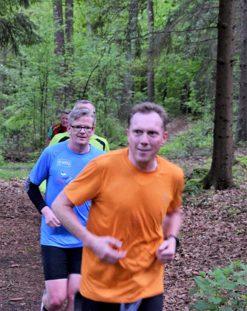 Läufer in sportlicher Kleidung joggen durch einen bewaldeten Pfad, umgeben von grünen Bäumen und frischer Natur.