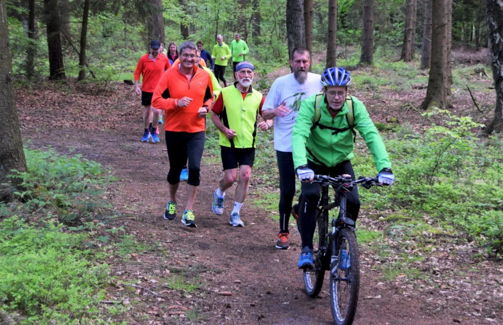Gruppe von Läufern und einem Radfahrer auf einem Waldweg, umgeben von grüner Natur, während sie aktiv Sport treiben. Die Teilnehmer tragen helle Sportkleidung und zeigen Freude an der Bewegung im Freien.