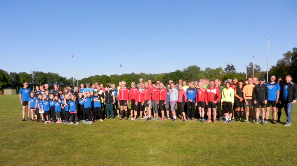 Gruppenfoto von Sportlern und Kindern in blauen und roten Trikots auf einem grünen Sportplatz unter klarem Himmel, ideal für eine Laufveranstaltung oder ein Sportfest.