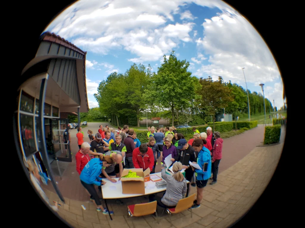 Gruppenevent im Freien mit Teilnehmern, die sich an einem Tisch anmelden. Sportler in bunten Trikots stehen vor einem Gebäude, umgeben von Bäumen und einer klaren Himmel.