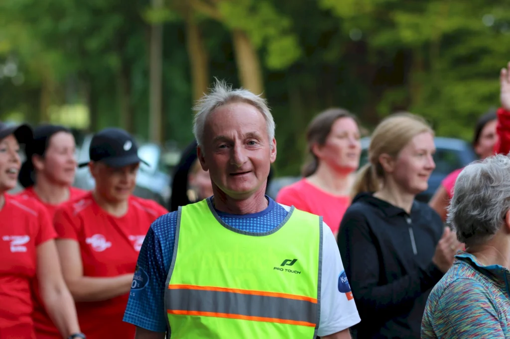 Mann in leuchtender Weste lächelt während einer Laufveranstaltung, umgeben von Teilnehmerinnen in roten Shirts, die aktiv mitmachen. Hintergrund zeigt eine grüne Umgebung.