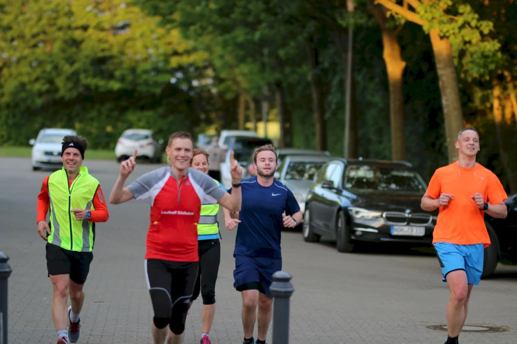 Gruppe von fünf Joggern in sportlicher Kleidung, die auf einer Straße laufen, umgeben von Bäumen und parkenden Autos. Der Fokus liegt auf der Freude am Laufen und der Gemeinschaft. Ideal für Themen wie Fitness, Lauftraining und Outdoor-Aktivitäten.