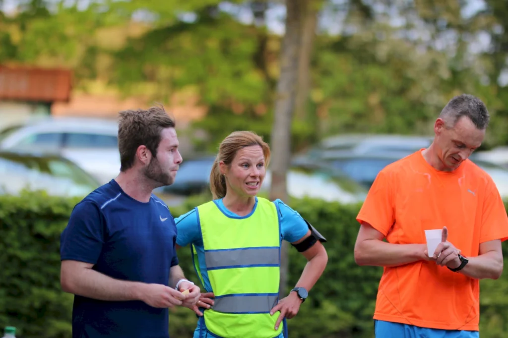 Drei Sportler in Laufbekleidung stehen im Freien zusammen. Die Frau in der Mitte trägt eine leuchtend grüne Weste und wirkt engagiert im Gespräch. Die Männer auf beiden Seiten scheinen auf sie zu hören, während sie sich auf eine sportliche Aktivität vorbereiten. Im Hintergrund sind verschwommen Autos und Bäume zu sehen.