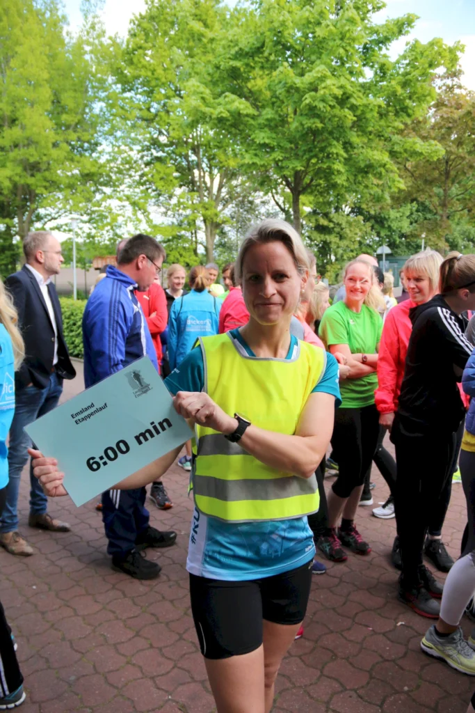 Teilnehmerin beim Emsland Etappenlauf hält ein Schild mit der Zeitangabe 6:00 min, umgeben von anderen Läufern in sportlicher Kleidung und einer grünen Umgebung.