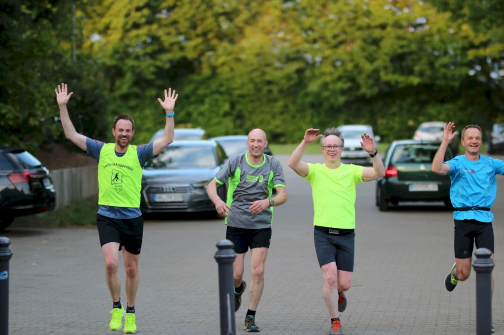 Fünf Männer in Sportkleidung laufen fröhlich auf einer Straße, heben die Hände und zeigen Begeisterung beim Laufen. Die Bäume im Hintergrund und parkende Autos schaffen eine lebendige Atmosphäre. Ideal für Themen wie Laufgemeinschaften und Fitnessaktivitäten.