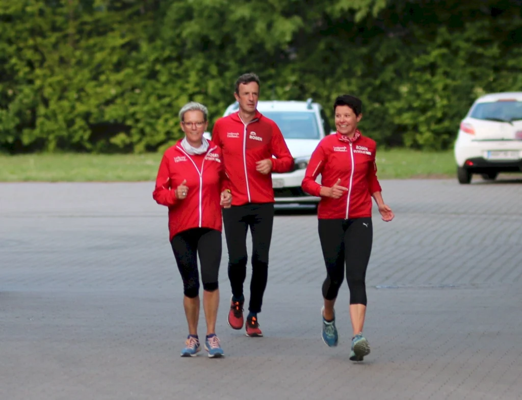 Drei Läufer in roten Jacken joggen auf einem gepflasterten Weg, umgeben von grüner Vegetation.