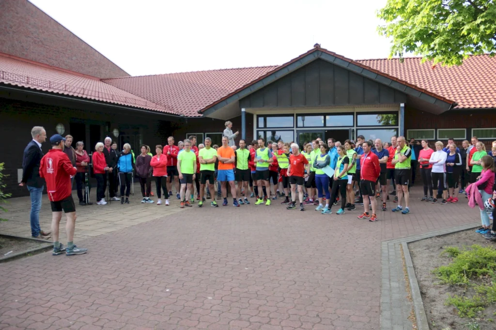 Gruppensportveranstaltung mit Läufern in bunten Trainingsanzügen vor einem Veranstaltungsgebäude, während eine Ansprache gehalten wird.