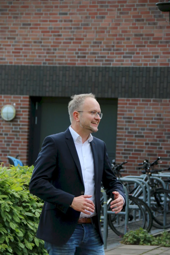 Ein lächelnder Mann in einem Anzug steht vor einer Backsteinmauer und gestikuliert. Im Hintergrund sind Fahrräder und grüne Pflanzen sichtbar.