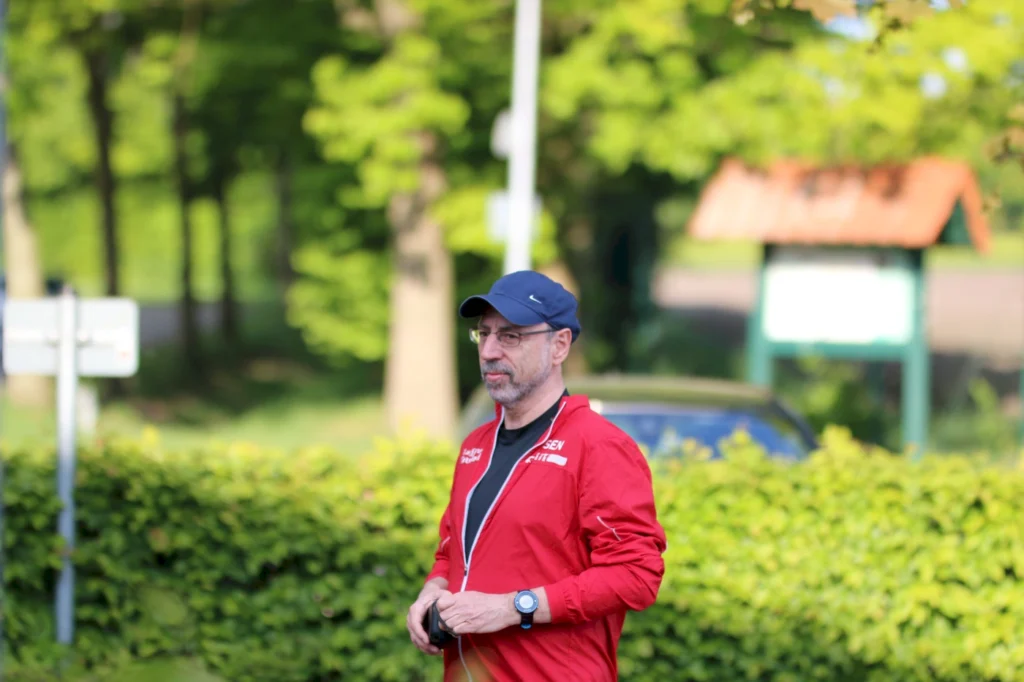 Mann in roter Sportjacke und blauer Cap steht in einem parkähnlichen Bereich, umgeben von grünen Bäumen und Sträuchern. Er hält ein Gerät in der Hand und sieht nachdenklich aus.