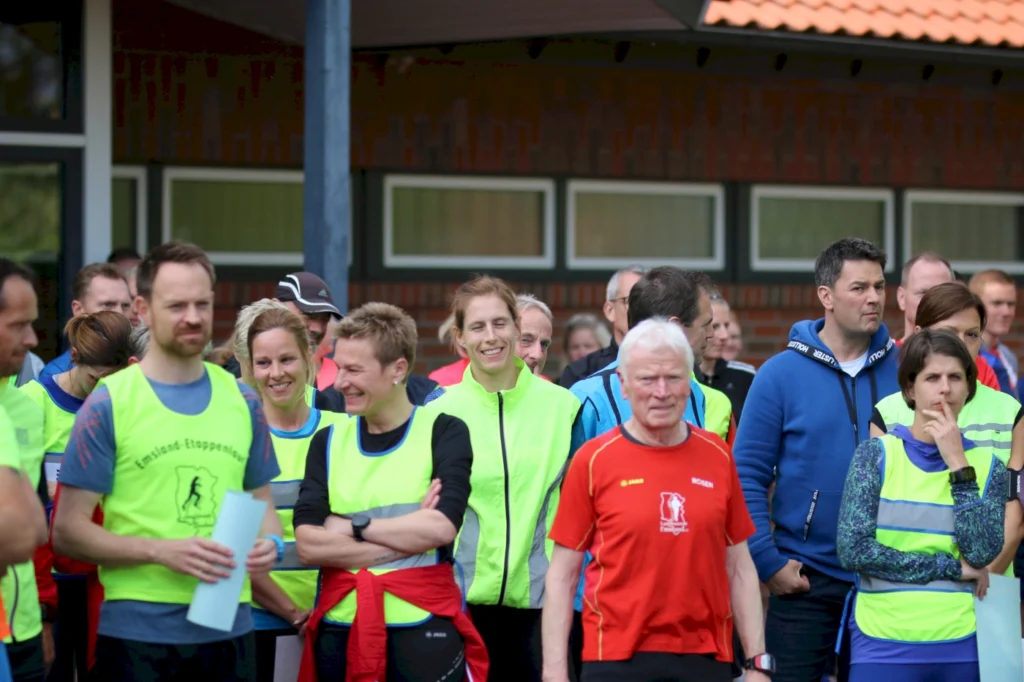 Gruppierung von Läufern in leuchtenden Westen, die sich vor einem Wettkampf versammeln. Die Teilnehmer lächeln und wirken entspannt, während sie auf den Start warten.