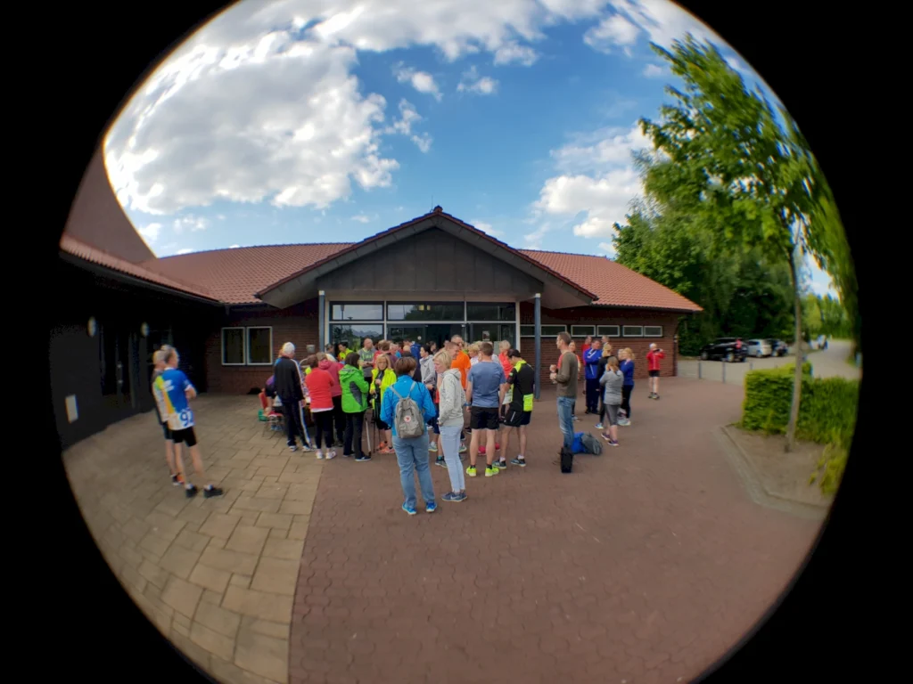 Gruppierung von Sportlern vor einem Veranstaltungsgebäude, die sich auf einen Wettkampf vorbereiten. Die Teilnehmer tragen bunte Sportkleidung und stehen auf einem gepflasterten Platz bei sonnigem Wetter.