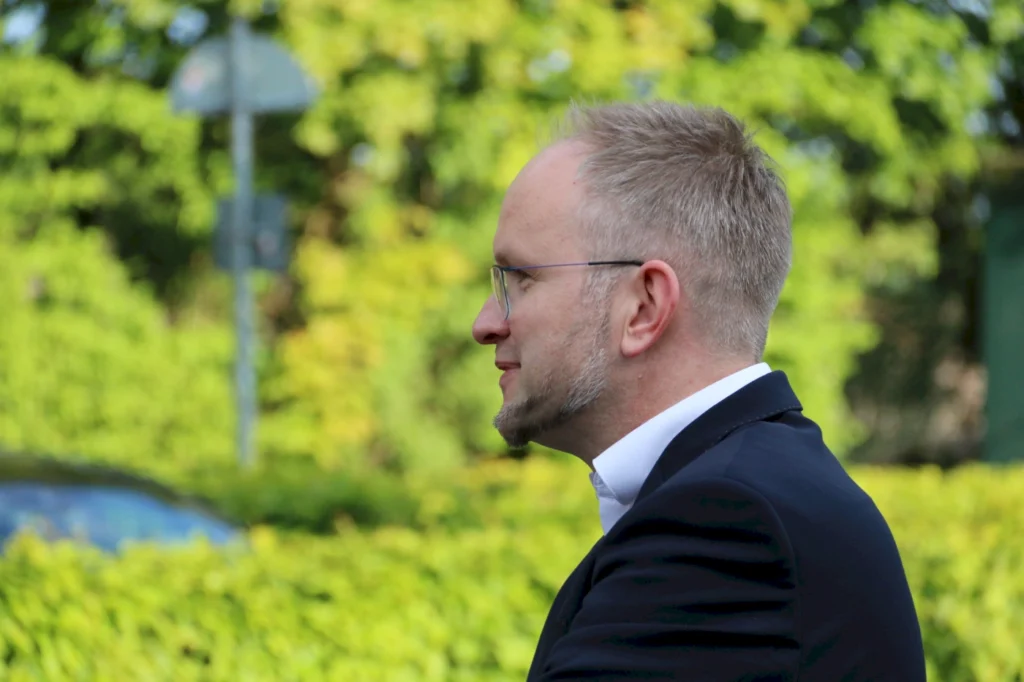 Mann im Anzug mit Brille, der im Profil steht, umgeben von grünem Hintergrund und Bäumen.