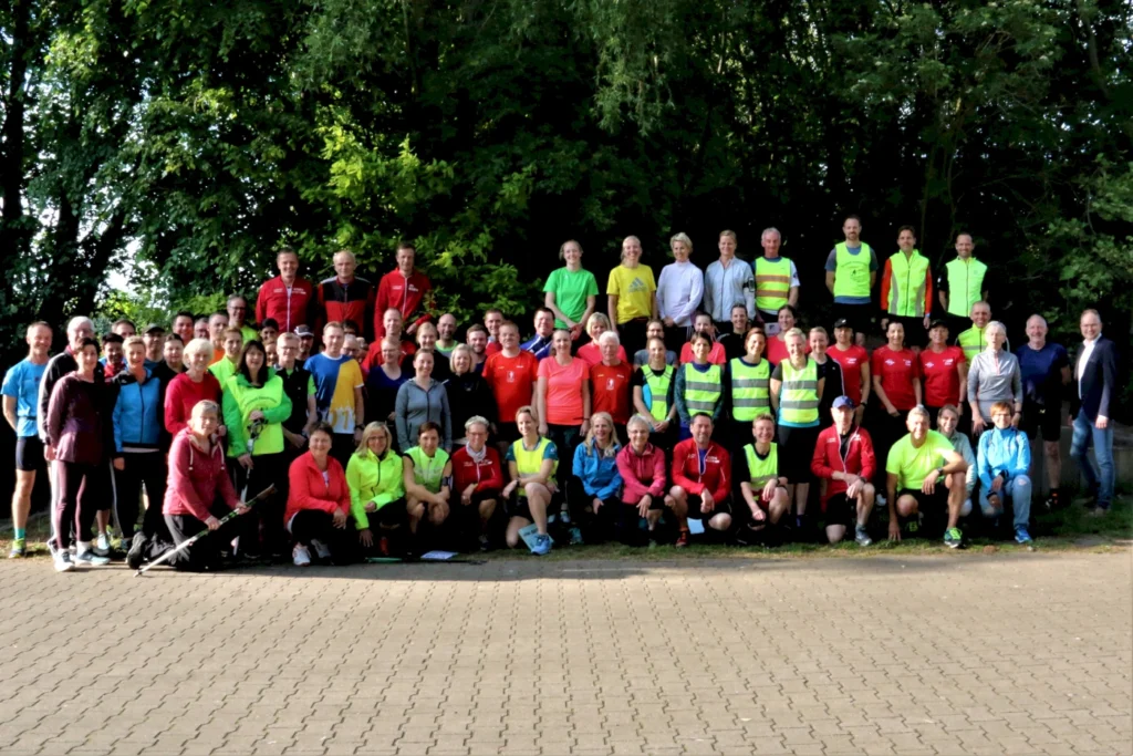 Gruppenfoto von Teilnehmern eines Lauf-Events, die in bunten Sportoutfits posieren. Die Gruppe besteht aus Männern und Frauen unterschiedlichen Alters und steht vor einer grünen Baumkulisse. Ideal für Themen wie Sportgemeinschaften, Laufveranstaltungen und Fitness-Events.
