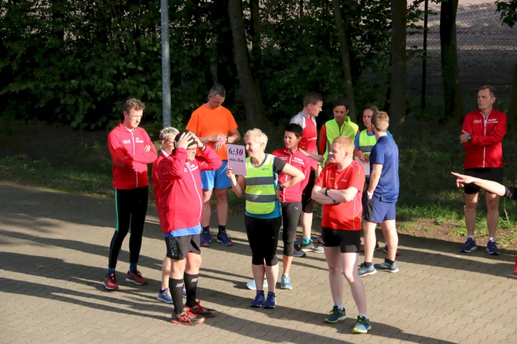 Gruppentraining von Läufern in Sportkleidung, die auf einer gepflasterten Fläche stehen. Einige Teilnehmer tragen rote Jacken, während andere in bunten T-Shirts gekleidet sind. Eine Person hält ein Schild mit der Aufschrift "6:30 min/km". Die Szene spielt sich im Freien vor einer begrünten Kulisse ab.