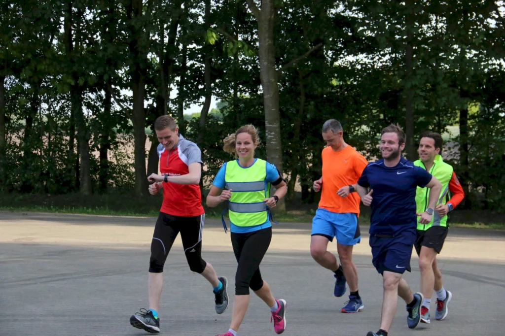 Gruppe von fünf Läufern, die in einem Park joggen, tragen farbenfrohe Sportbekleidung und lachen. Ideal für Fitness- und Laufveranstaltungen.