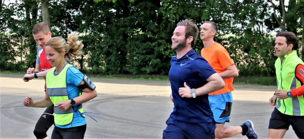 Gruppe von fünf Läufern, die auf einer Straße joggen, umgeben von Bäumen. Die Teilnehmer tragen unterschiedliche Laufbekleidung in lebhaften Farben und zeigen Freude beim Laufen.