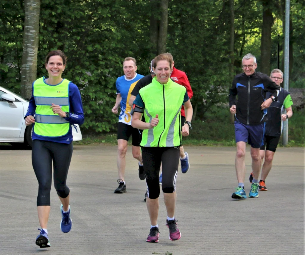 Gruppe von fünf Joggern, die in einem Park lächeln und aktiv laufen, während sie sportliche Kleidung tragen. Im Hintergrund ist ein Auto und grüne Bäume sichtbar.