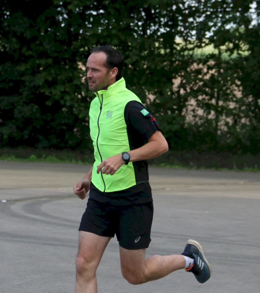 Läufer in leuchtend grünem Sportjacke und Shorts, der auf einer asphaltieren Strecke joggt. Die Szene zeigt eine natürliche Umgebung mit Bäumen im Hintergrund, ideal für Lauftraining und Fitnessaktivitäten.