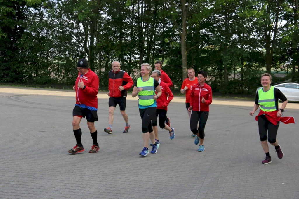Gruppe von Läufern in roten Trainingsjacken und leuchtenden Westen, die auf einem gepflasterten Weg joggen, umgeben von Bäumen. Ideal für Fitness- und Laufenthusiasten.
