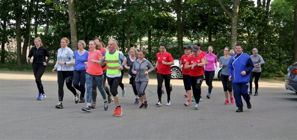 Gruppe von Läufern, darunter Männer und Frauen, die in Sportbekleidung joggen. Die Szene spielt sich im Freien ab, umgeben von Bäumen. Einige Teilnehmer tragen farbige T-Shirts, während andere in neutraleren Farben gekleidet sind.