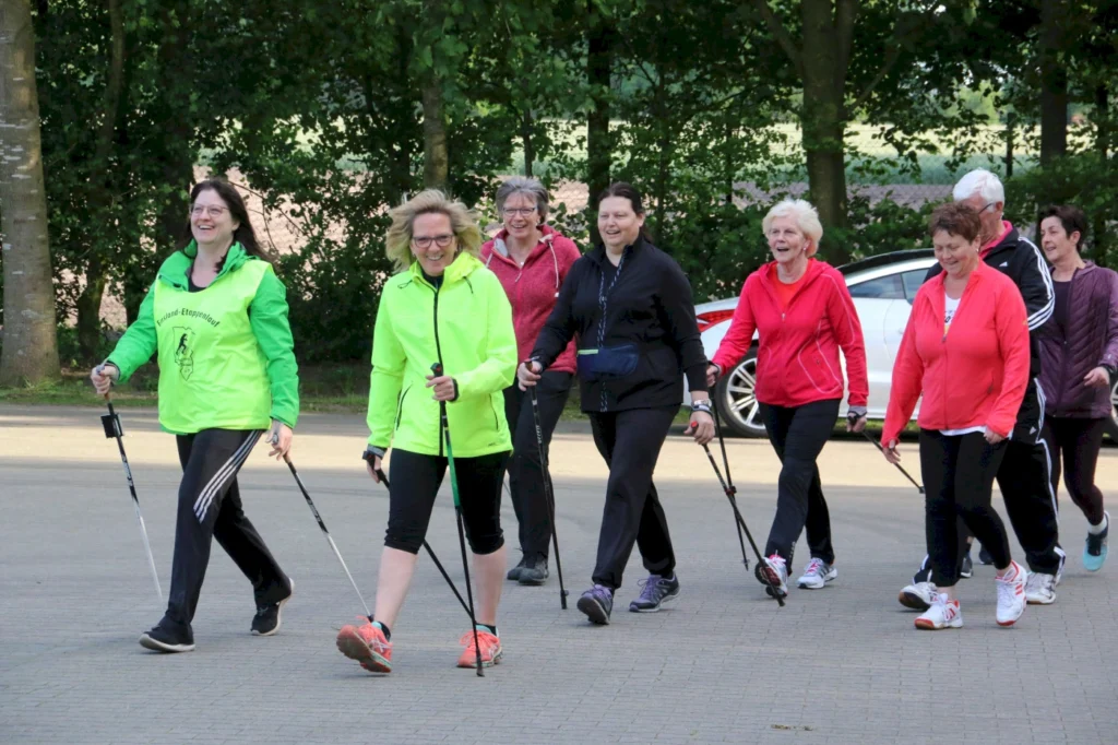 Gruppe von Frauen beim Nordic Walking in sportlicher Kleidung, lächelnd und mit Walkingstöcken, umgeben von Bäumen und einem Parkplatz.
