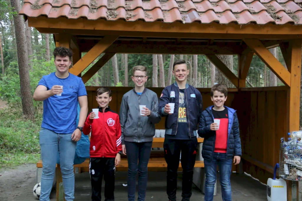Gruppe von fünf Jugendlichen, die in einem Wald unter einem Holzpavillon stehen und Pappbecher mit Getränken halten, umgeben von Bäumen und Natur.