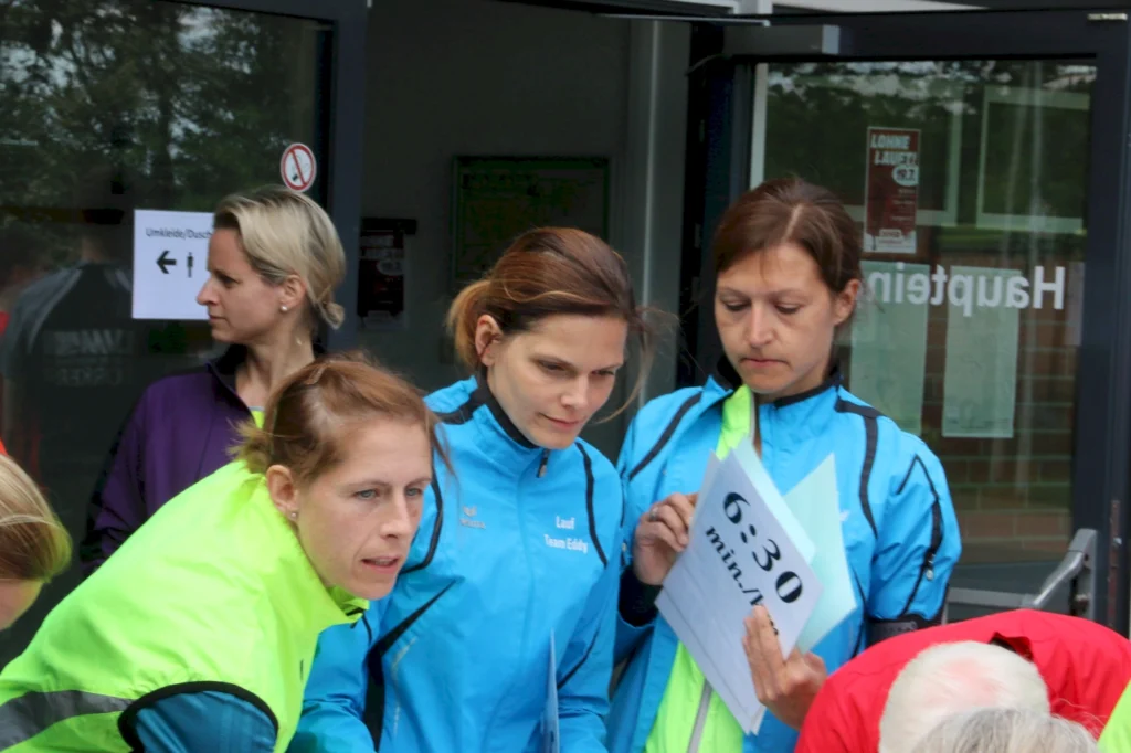 Drei Frauen in Laufbekleidung stehen vor einem Gebäude und schauen auf Informationsblätter. Eine Frau hält ein Blatt mit der Aufschrift "6:30 min/km". Im Hintergrund sind weitere Personen und ein Hinweis auf Umkleide/Duschen sichtbar.