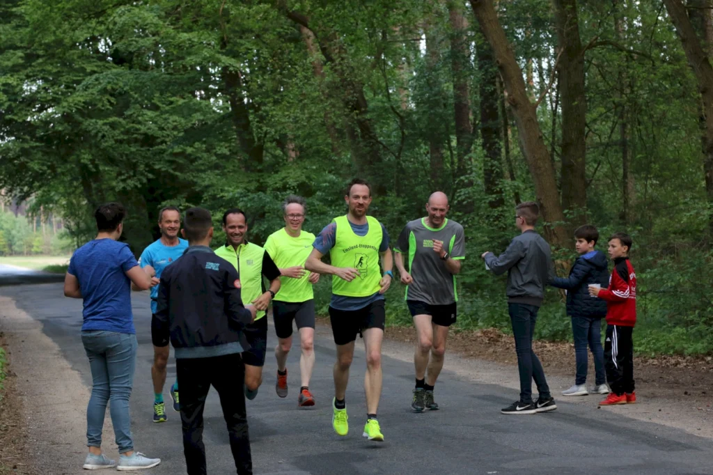 Gruppe von Läufern in leuchtenden Shirts, die auf einem Waldweg joggen, während Zuschauer am Rand stehen und anfeuern. Die Szene zeigt eine aktive Gemeinschaft und die Freude am Laufen in der Natur.