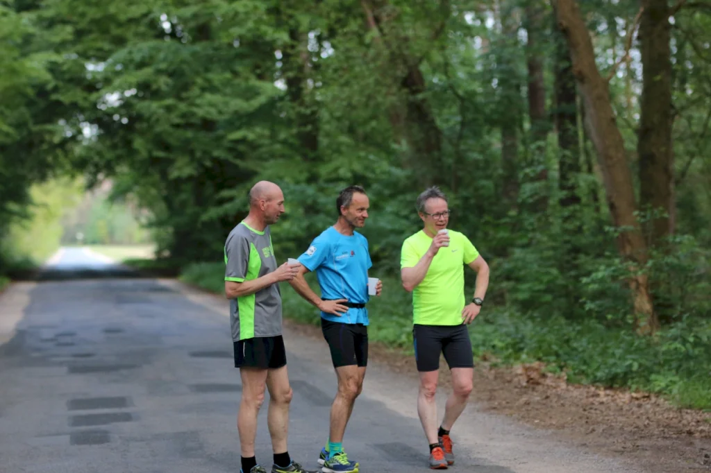 Drei Männer in Sportkleidung stehen auf einer ruhigen Straße im Wald und unterhalten sich nach dem Laufen, während sie Getränke halten.