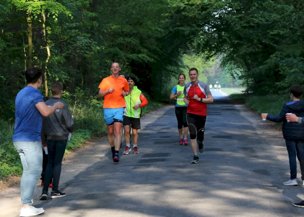 Läufer in sportlicher Kleidung joggen auf einem Waldweg, während Zuschauer am Rand stehen und sie anfeuern. Die Szene zeigt eine fröhliche Laufveranstaltung in der Natur.