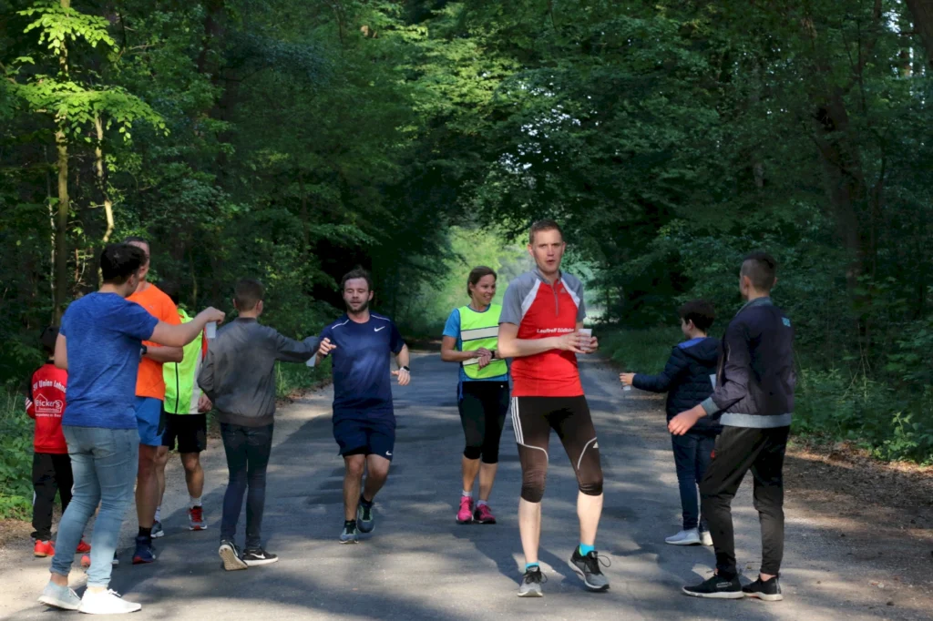 Gruppe von Menschen beim Laufen auf einem Waldweg, umgeben von Bäumen, mit verschiedenen Sportoutfits und einem Teilnehmer, der ein Getränk hält.