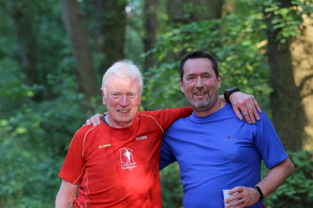 Zwei Männer stehen lächelnd in einem Wald, der älterer Mann trägt ein rotes T-Shirt mit dem Schriftzug "Rosen", der jüngere ein blaues T-Shirt. Beide haben einen freundlichen Gesichtsausdruck und scheinen eine gute Zeit zu haben.