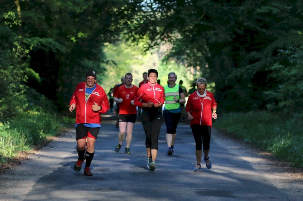 Gruppe von Läufern in roten Jacken joggt auf einem Waldweg, umgeben von grüner Natur. Ideal für Fitness- und Laufveranstaltungen.