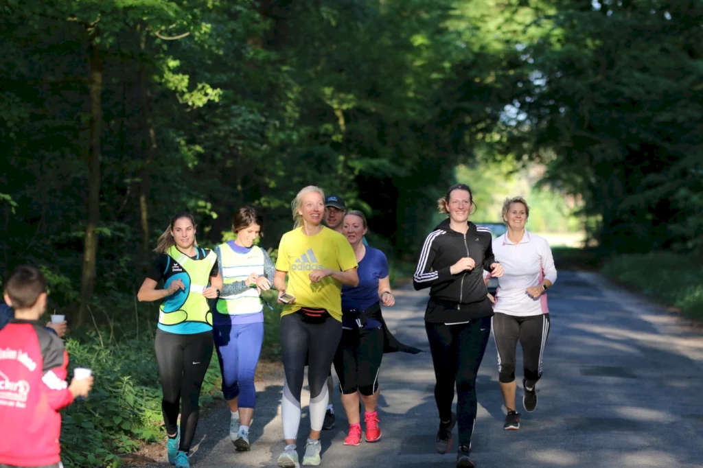 Gruppe von Frauen joggend auf einem Waldweg, während sie sich unterhalten und Spaß haben. Sonniger Tag mit grüner Umgebung, ideal für Outdoor-Sport.