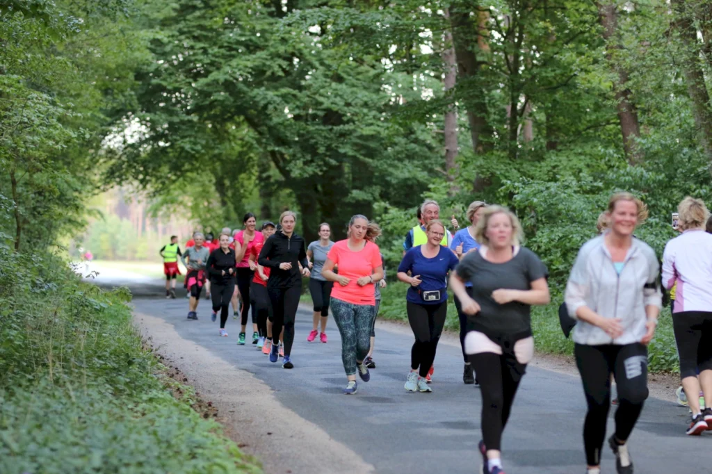 Gruppe von Frauen, die in einem Waldgebiet joggen, umgeben von grünen Bäumen und einer ruhigen Straße, ideal für Outdoor-Sport und Fitnessaktivitäten.