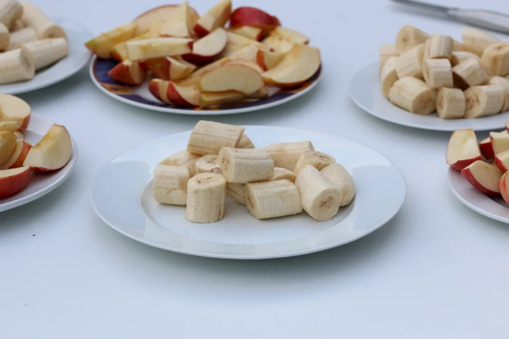 Auf einem weißen Tisch liegen mehrere Teller mit geschnittenen Äpfeln und Bananenstücken. Die Bananen sind in gleichmäßige Scheiben geschnitten und auf einem Teller angerichtet, während die Äpfel in Viertel geschnitten sind und auf verschiedenen Tellern präsentiert werden.