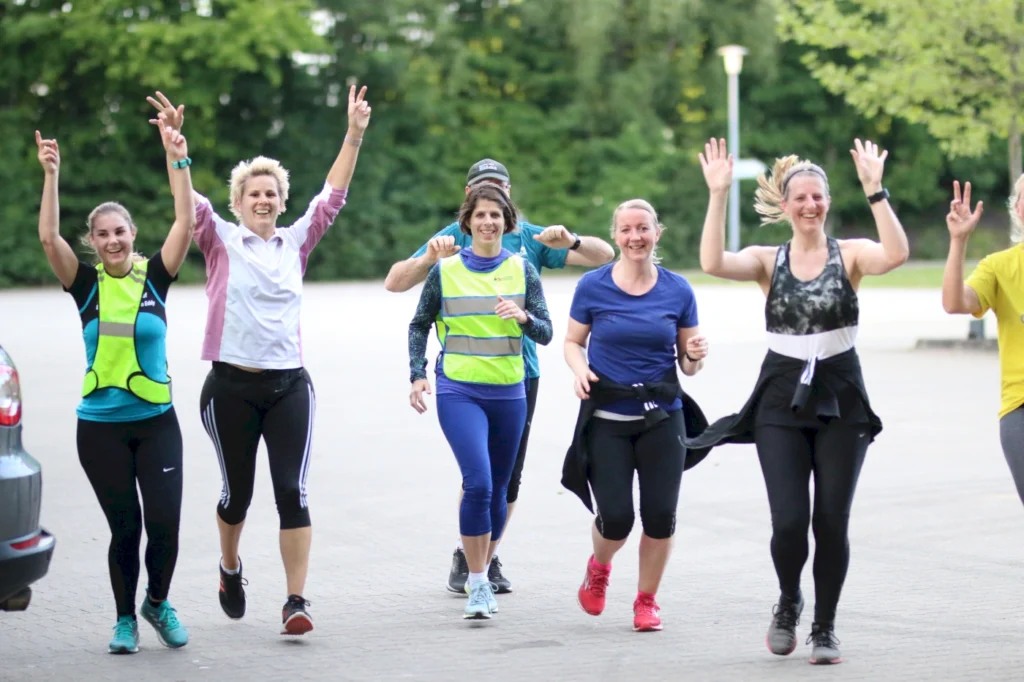 Gruppe von Frauen beim Laufen, die fröhlich die Hände heben und sich gegenseitig anfeuern, in einer grünen Umgebung. Sie tragen Sportbekleidung und reflektierende Westen, was auf ein gemeinsames Lauftraining hinweist.