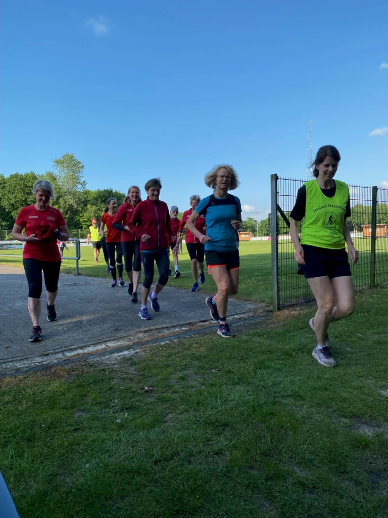 Gruppe von Frauen in Sportbekleidung beim Laufen auf einem Platz im Freien, umgeben von Grünflächen und blauem Himmel.