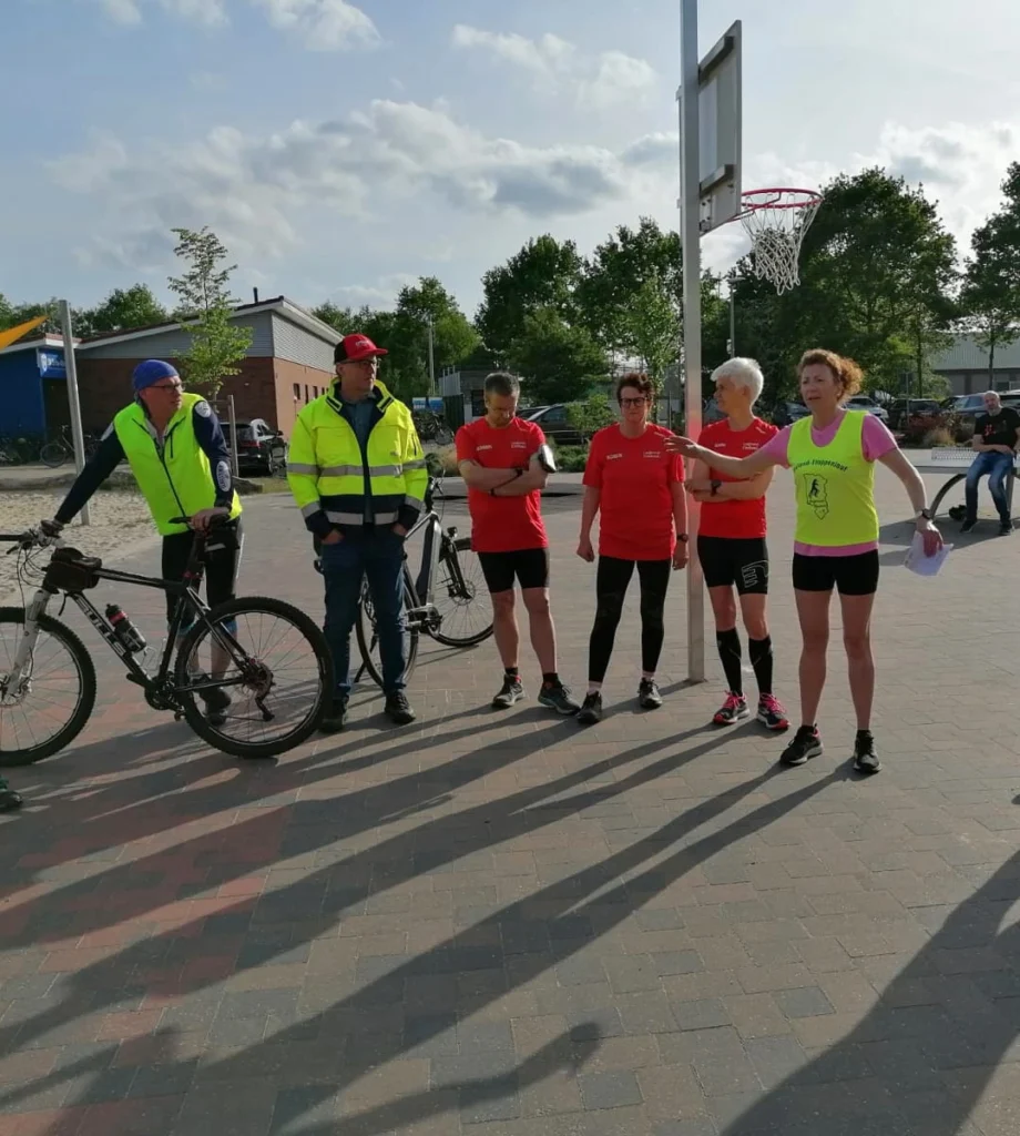 Gruppensitzung von Sportlern und Helfern im Freien, während eines Events. Einige Personen tragen rote Sportbekleidung, andere gelbe Warnwesten. Im Hintergrund sind Fahrräder und ein Basketballkorb zu sehen.
