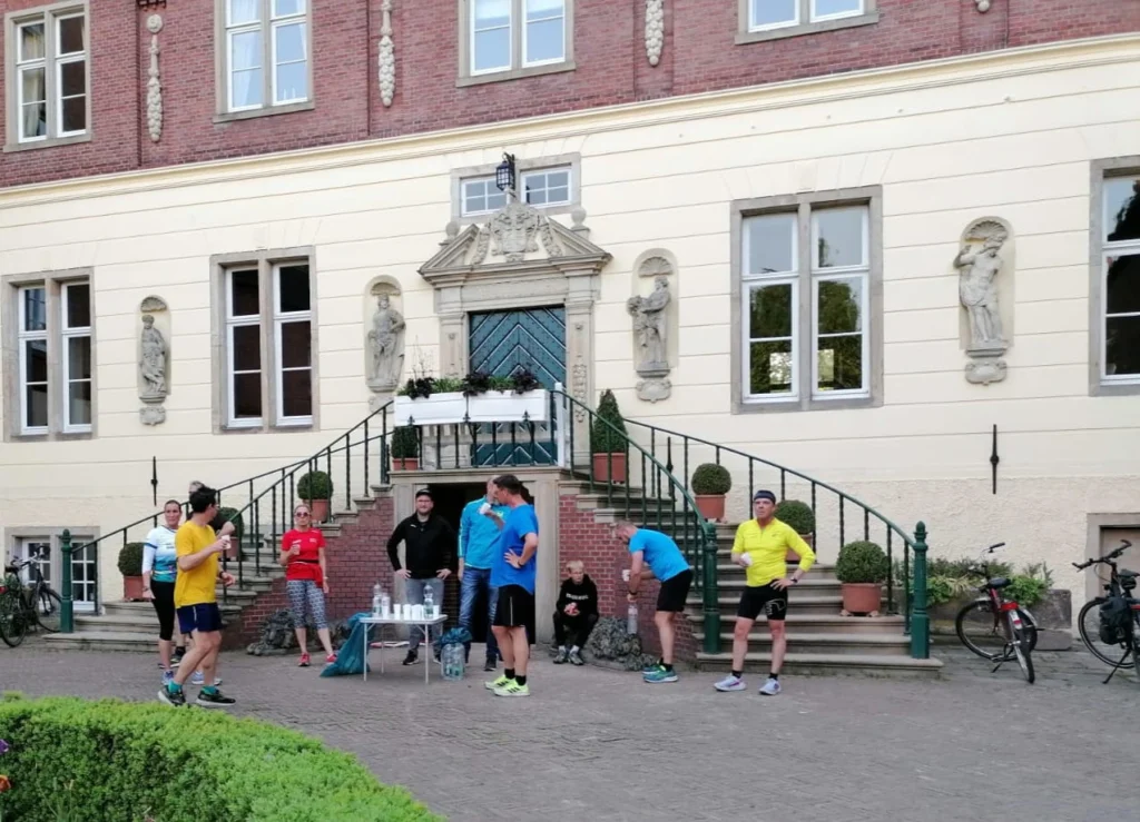 Gruppensportveranstaltung vor einem historischen Gebäude, mit Teilnehmern in Sportkleidung, die sich auf einen Lauf vorbereiten und Wasserflaschen auf einem Tisch neben ihnen.