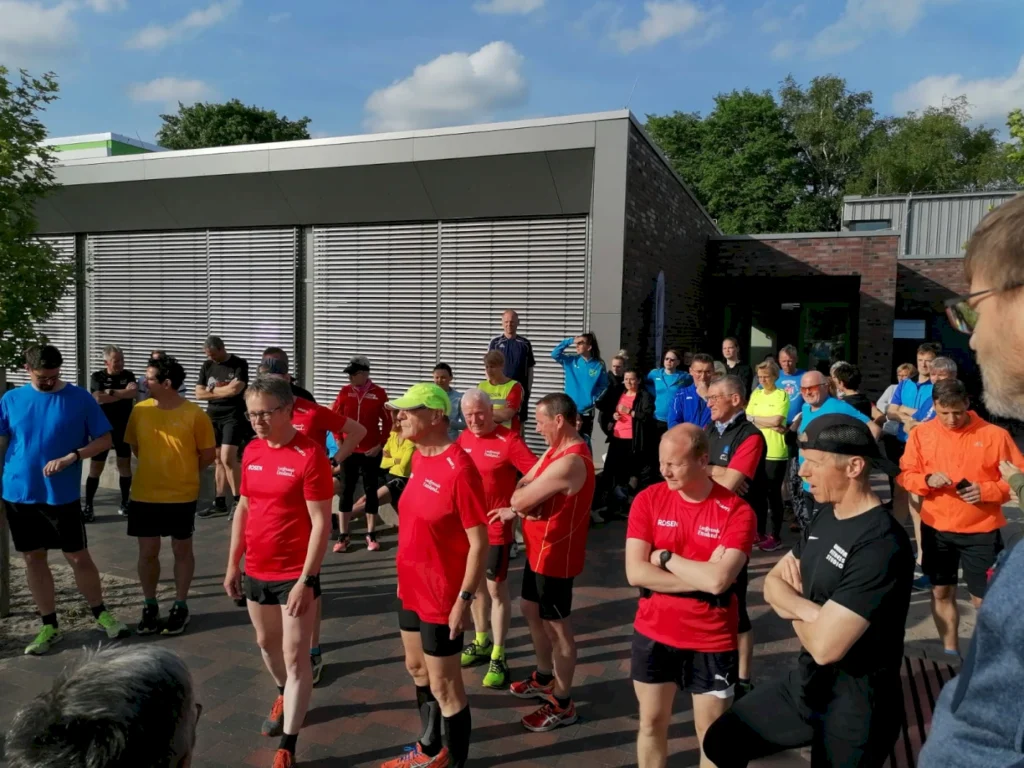 Gruppierung von Läufern in bunten Trikots vor einem modernen Gebäude, bereit für einen Lauf oder Wettkampf. Die Szene zeigt Teilnehmer in verschiedenen Altersgruppen, die sich auf den Start vorbereiten, bei sonnigem Wetter.