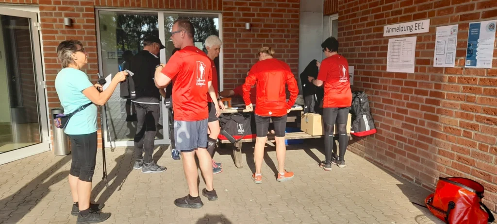 Teilnehmer einer Veranstaltung stehen an einem Anmeldetisch im Freien, während sie sich auf die Aktivität vorbereiten. Einige Personen tragen rote T-Shirts mit einem Logo, andere haben Wanderstöcke und Notizen in der Hand. Im Hintergrund sind Informationsblätter an der Wand angebracht.