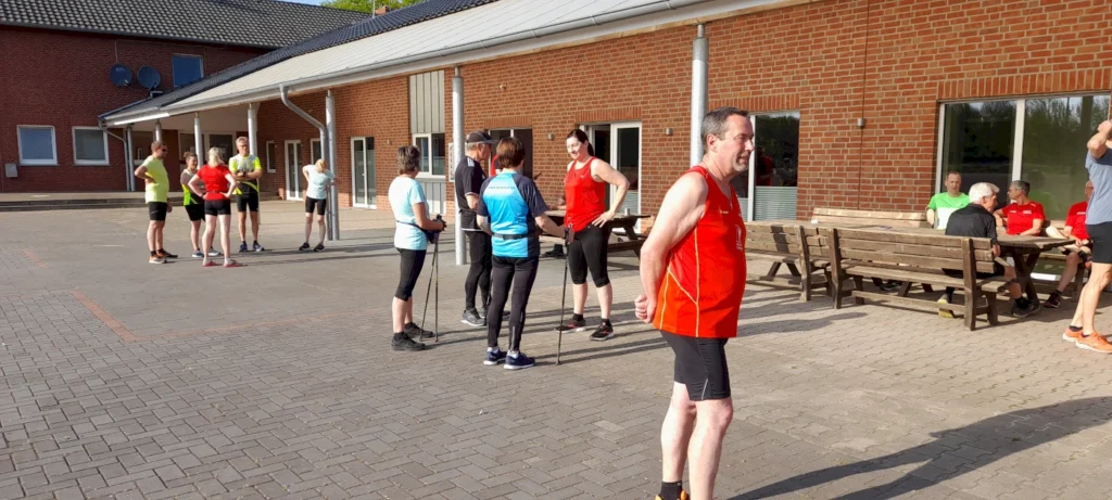 Gruppentraining im Freien mit Sportlern in bunten Trikots, die sich auf eine Nordic-Walking-Session vorbereiten. Im Hintergrund ist ein Sportgebäude und Sitzgelegenheiten zu sehen.