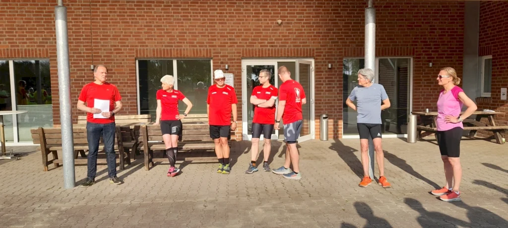 Gruppenszene mit sechs Läufern in roten T-Shirts, die vor einem Gebäude stehen. Eine Person hält ein Blatt Papier, während die anderen aufmerksam zuhören. Die Umgebung ist hell erleuchtet und zeigt eine entspannte Atmosphäre, ideal für eine Laufveranstaltung oder ein Training.