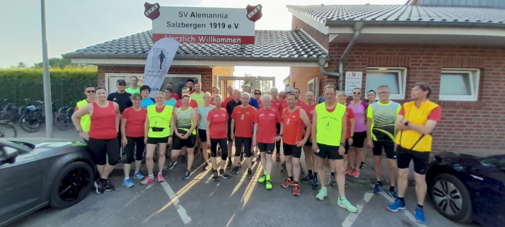 Gruppenfoto von Laufbegeisterten vor dem Vereinsgebäude des SV Alemannia Salzbergen, die sich für eine Laufveranstaltung versammeln. Die Teilnehmer tragen bunte Laufkleidung und stehen auf dem Parkplatz, während Fahrräder im Hintergrund abgestellt sind.