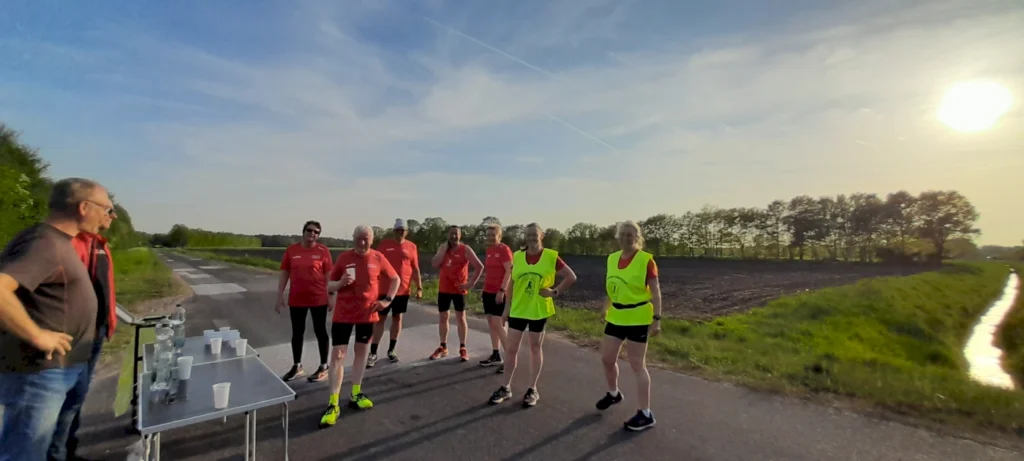 Gruppe von Läufern in roten Trikots und gelben Warnwesten, die an einer Strecke stehen, während ein Mann am Tisch mit Getränken bereitsteht. Die Szene findet bei sonnigem Wetter und klarem Himmel statt, umgeben von grüner Landschaft und Bäumen.